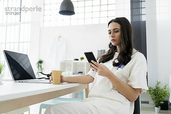 Junge Ärztin mit Smartphone am Schreibtisch in der Klinik
