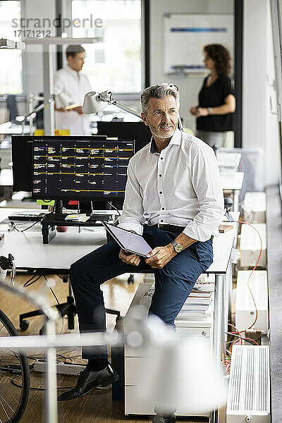 Geschäftsmann mit digitalem Tablet  der wegschaut  während er mit einem Kollegen im Hintergrund in einem Großraumbüro auf dem Schreibtisch sitzt