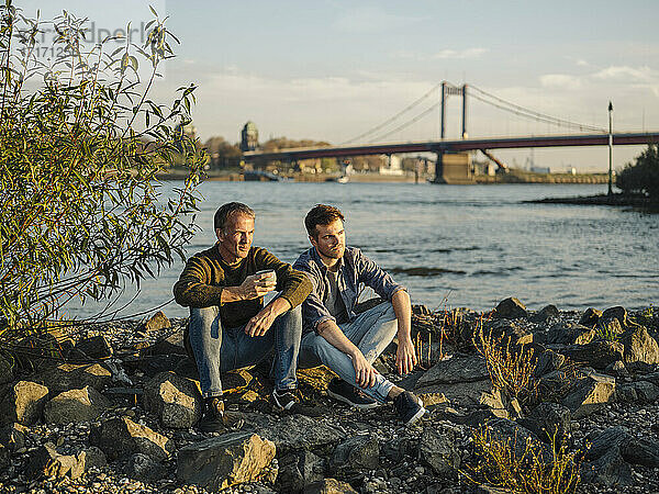 Father and son sitting on rock against river at evening