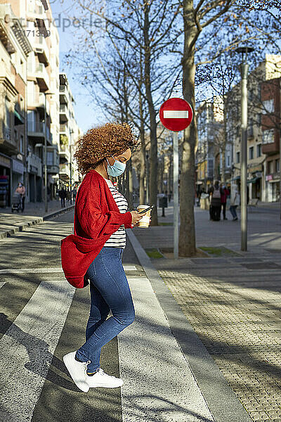 Afro woman wearing protective face mask using mobile phone while walking on road in city