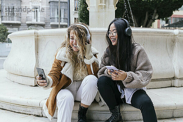 Laughing female friends taking selfie wearing headphones while sitting against fountain
