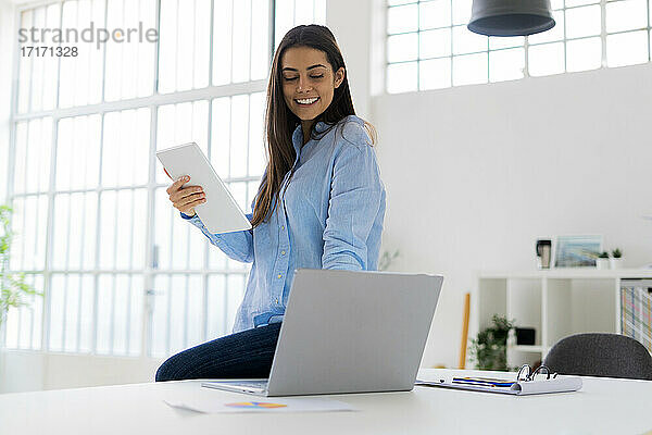Weibliche Fachkraft hält digitales Tablet  während sie im Büro am Laptop arbeitet