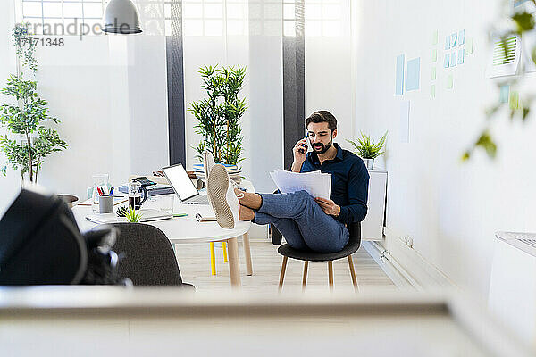 Young businessman discussing on smart phone while sitting with feet up at desk in office