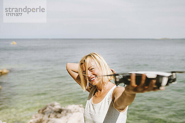 Lächelnde Frau  die eine Drohne hält  während sie am Meer steht