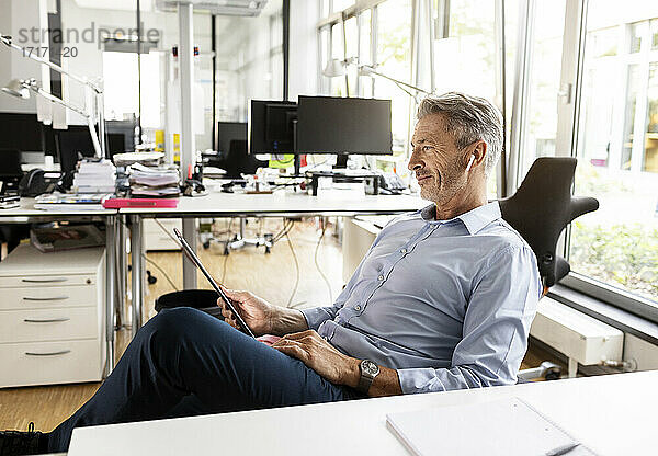 Geschäftsmann benutzt ein digitales Tablet  während er sich auf einem Stuhl in einem Großraumbüro entspannt