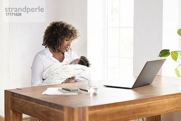 Mutter sieht schlafendes Baby an  während sie am Tisch im Büro zu Hause sitzt