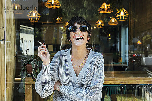 Junge Frau mit Sonnenbrille raucht eine Zigarette  während sie am Fenster eines Restaurants steht
