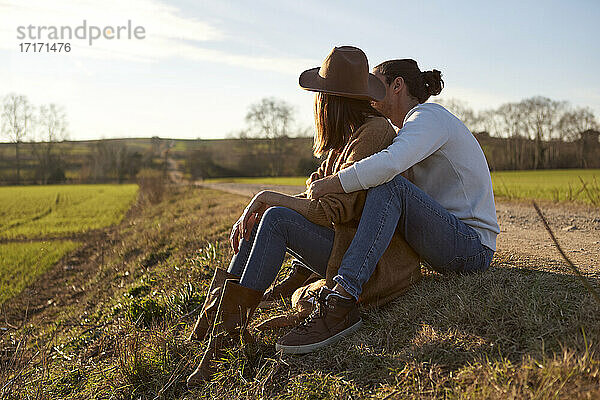 Boyfriend embracing girlfriend from behind while sitting on field during sunset