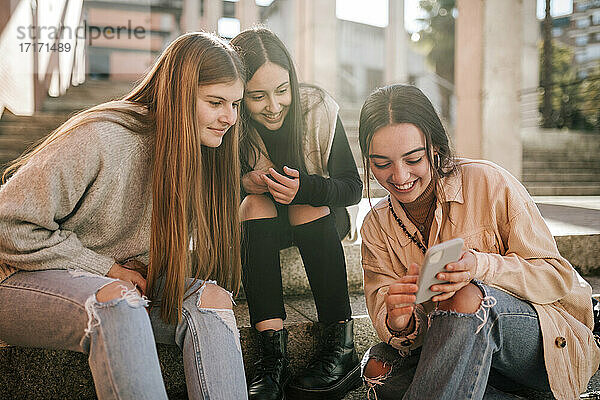 Lächelndes Teenager-Mädchen  das Freunden das Handy zeigt  während es auf einer Treppe in der Stadt sitzt