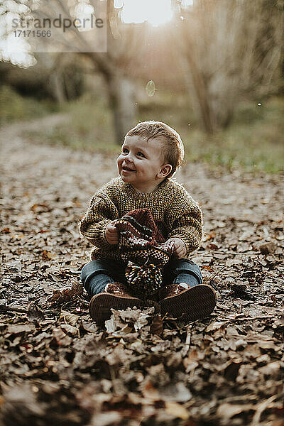 Fröhliches Kleinkind mit Strickmütze auf trockenem Herbstlaub sitzend und in den Wald schauend