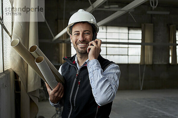 Lächelnder Immobilienentwickler mit Bauplänen  der über ein Mobiltelefon in einem Gebäude spricht