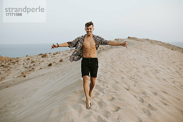 Fröhlicher junger Mann  der das Shaka-Zeichen macht  während er auf Sanddünen in Almeria  Tabernas-Wüste  Spanien  spazieren geht