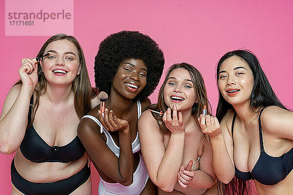 Fröhliche multiethnische weibliche Modelle in Unterwäsche  die Schönheitsprodukte vor einem rosa Hintergrund halten