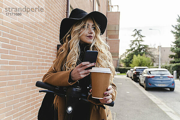 Young woman with electric push scooter holding reusable coffee cup while looking away