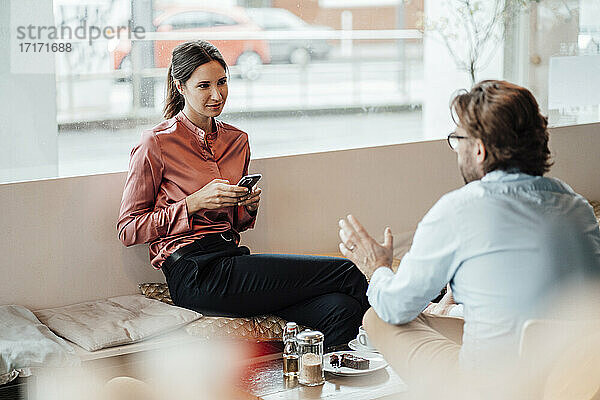 Geschäftsfrau hält ihr Smartphone in der Hand  während sie mit einem männlichen Kollegen im Café diskutiert