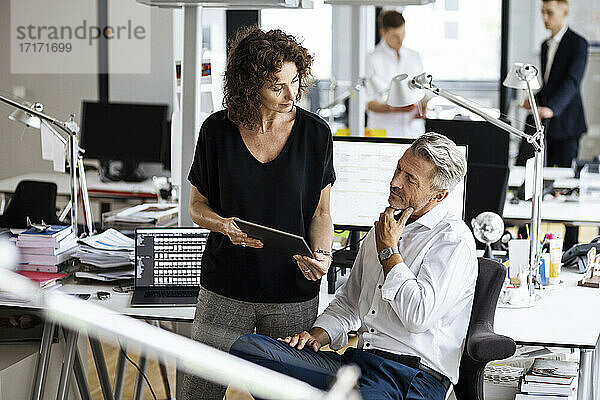 Ältere Geschäftsleute verwenden ein digitales Tablet  während sie im Hintergrund mit Kollegen in einem Großraumbüro arbeiten