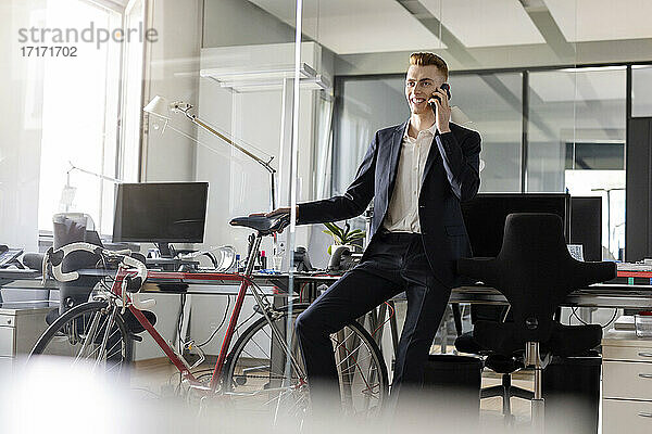 Männlicher Unternehmer mit Fahrrad  der lächelt  während er in einem Großraumbüro sitzt und mit einem Mobiltelefon spricht