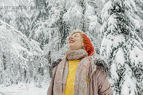Lächelnde Frau genießt den Schneefall  während sie im Wald steht