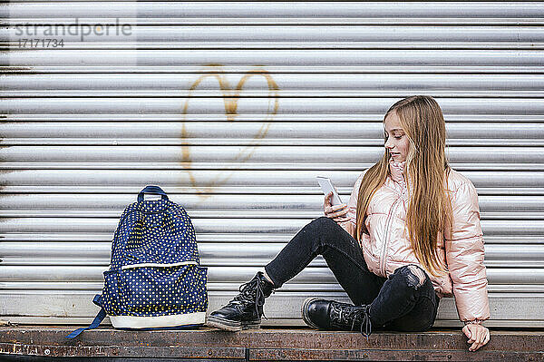 Girl with long brown hair looking at mobile phone while sitting against shutter