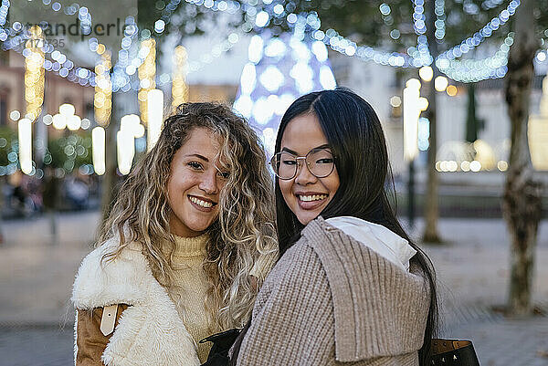 Young female friends smiling against illuminated Christmas decoration at dusk