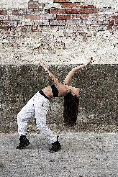 Flexible female dancer bending backwards while dancing against old brick wall in factory