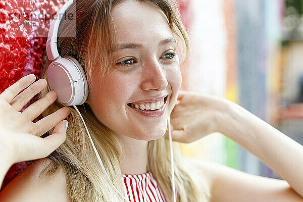 Lächelnde junge Frau  die wegschaut  während sie mit Kopfhörern an einer bunten Wand Musik hört