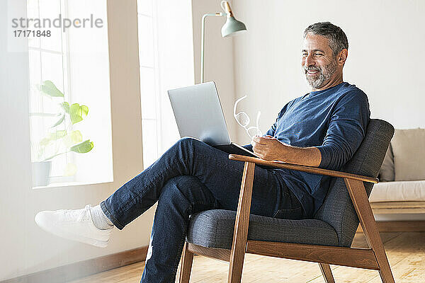 Lächelnder Geschäftsmann  der einen Laptop benutzt  während er zu Hause auf einem Sessel sitzt