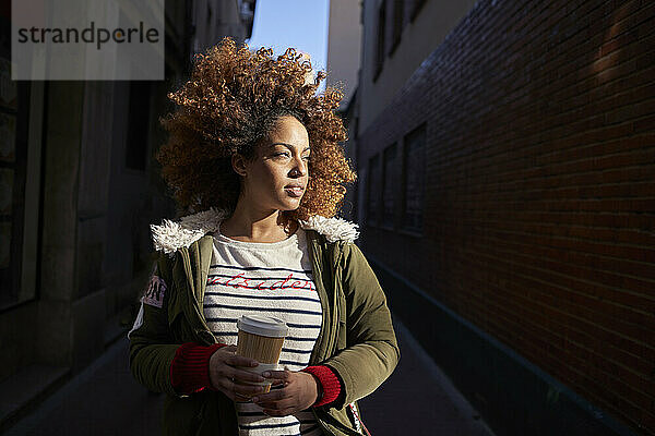 Young woman with disposable coffee cup looking away while standing on footpath