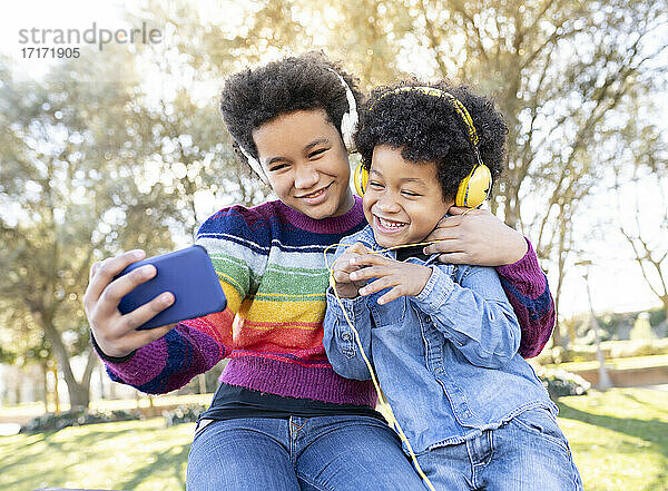 Geschwister mit Kopfhörern  die lächelnd ein Selfie mit dem Handy machen  während sie im Park sitzen