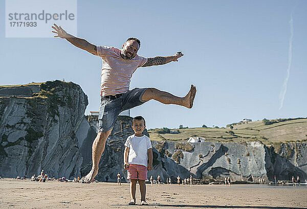 Cheerful father jumping over son at beach against sky