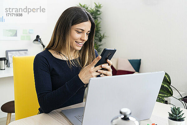 Lächelnder Freiberufler  der ein Mobiltelefon benutzt  während er im Wohnzimmer am Laptop sitzt