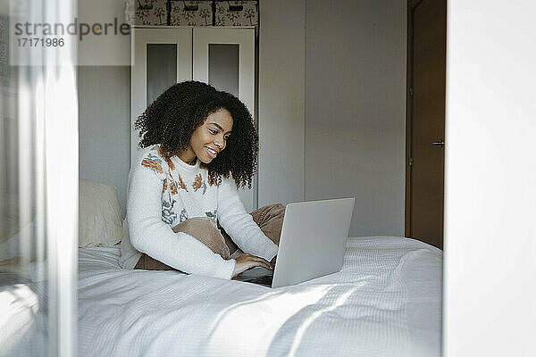 Lächelnde Frau  die einen Laptop benutzt  während sie zu Hause auf dem Bett sitzt