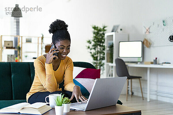 Smiling young woman talking on mobile phone while using laptop in living room