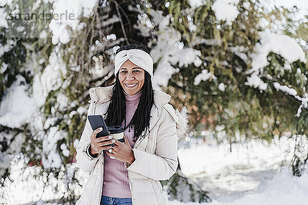 Lächelnde Frau mit Smartphone und Kaffeetasse im Park im Winter