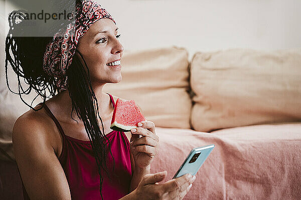 Frau mit Handy isst Wassermelone  während sie zu Hause sitzt