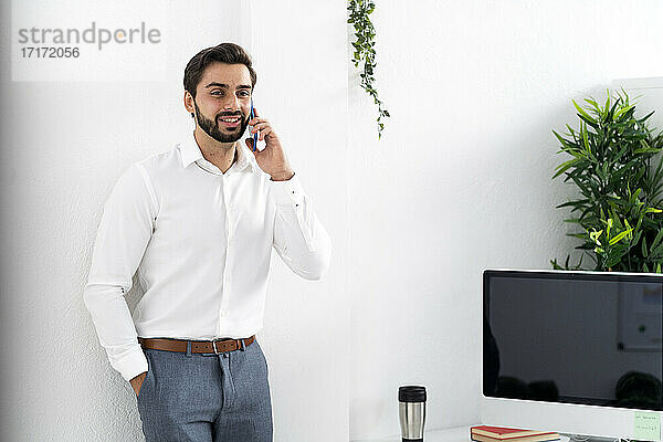 Männlicher Berufstätiger mit Händen in den Taschen bei einem Telefongespräch an einer weißen Wand am Arbeitsplatz