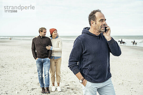 Porträt eines Mannes  der am Sandstrand steht und mit einem jungen Paar im Hintergrund über sein Smartphone spricht