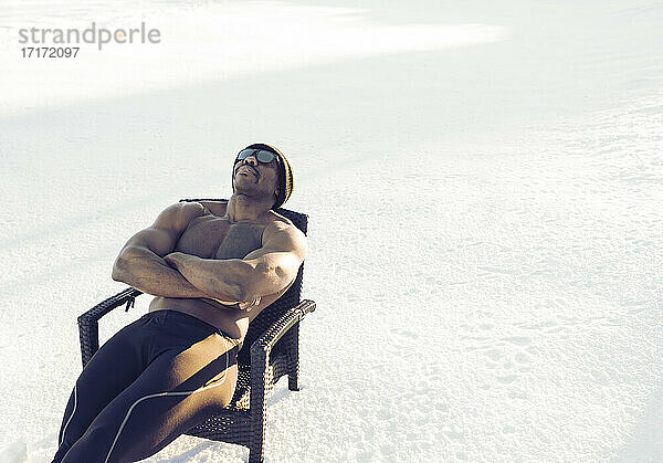 Mittlerer erwachsener Sportler mit Sonnenbrille  der sich ausruht  während er mit verschränkten Armen auf einem Stuhl im Schnee sitzt