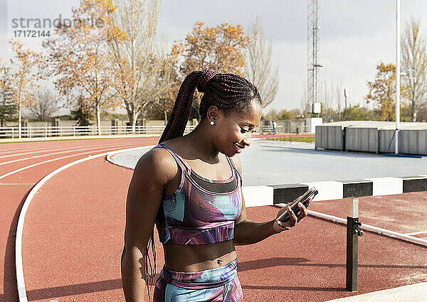 Lächelnde Sportlerin mit geflochtenen Haaren  die an einem sonnigen Tag ihr Smartphone auf der Rennstrecke benutzt