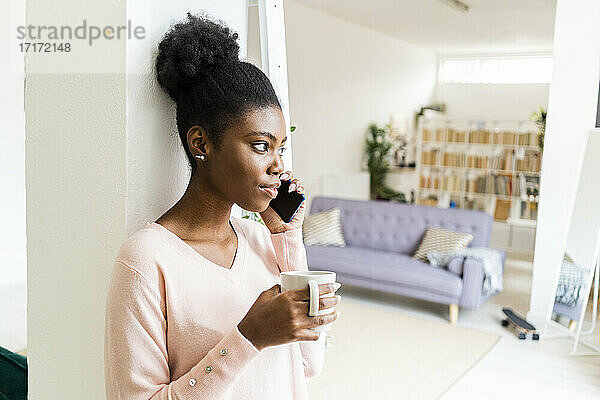 Junge Frau mit Kaffeetasse  die im Stehen zu Hause mit ihrem Handy telefoniert