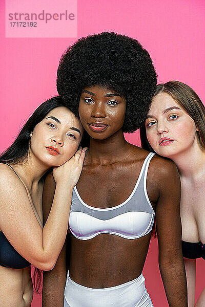 Multi-ethnische weibliche Modelle in Dessous lehnen sich an die Schulter einer Afro-gestylten Freundin vor einem rosa Hintergrund
