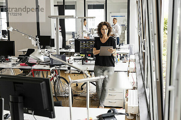Geschäftsfrau arbeitet an einem digitalen Tablet  während sie mit einem Kollegen im Hintergrund in einem Großraumbüro steht