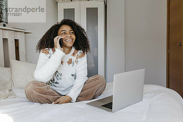Lächelnde Frau mit Laptop  die zu Hause sitzt und mit ihrem Handy telefoniert