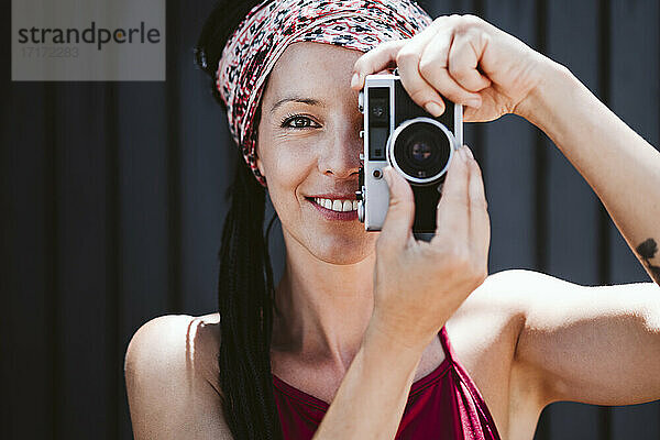 Lächelnde Frau  die durch die Kamera fotografiert  während sie an der Wand steht