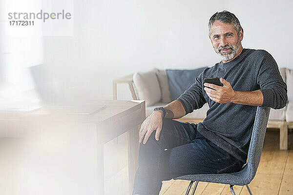 Selbstbewusster Geschäftsmann  der ein Mobiltelefon benutzt  während er im Büro zu Hause sitzt