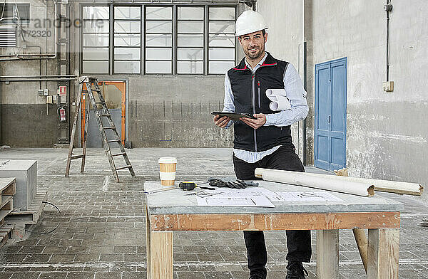 Lächelnder männlicher Architekt mit digitalem Tablet an einem Tisch auf einer Baustelle