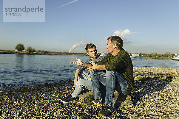 Father giving advice to son while sitting on rock at riverbank during sunny day
