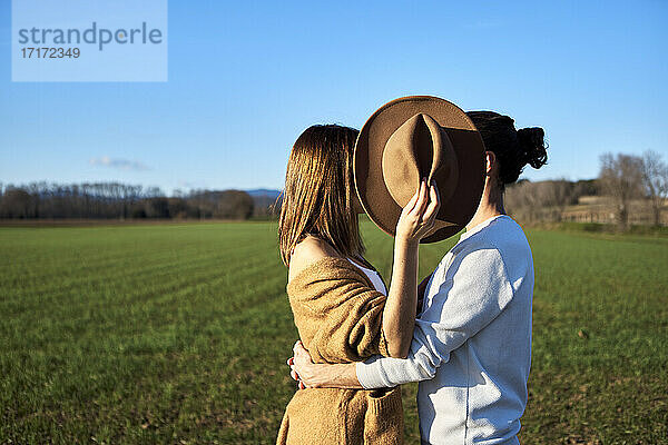 Heterosexual couple embracing while girlfriend covering faces with hat against clear blue sky