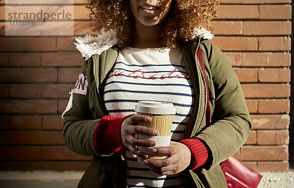 Woman wearing jacket holding disposable coffee cup while standing against brick wall