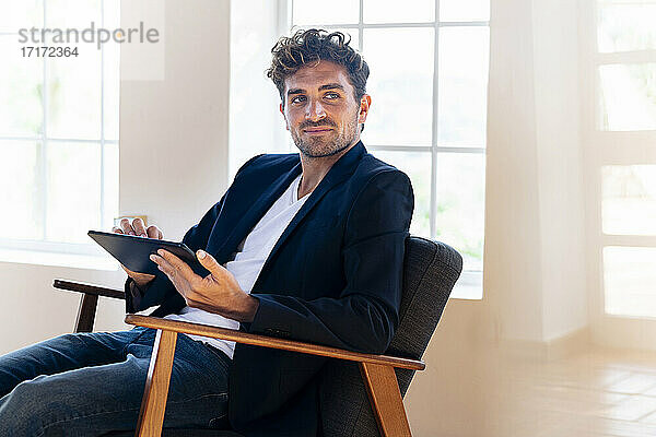 Geschäftsmann hält digitales Tablet  während er zu Hause auf einem Sessel sitzt
